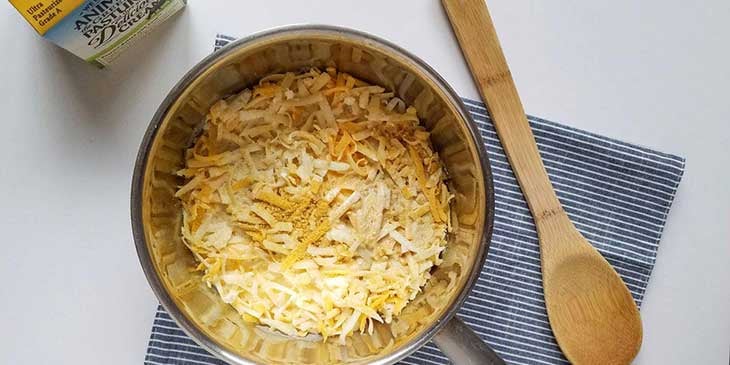 cheese sauce in a stainless steel saucepan next to a wooden spatula and a carton of heavy cream