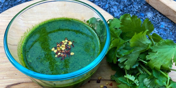 Keto Zhoug sauce plated on a round glass small bowl next to fresh cilantro leaves on top of a wooden cutting board